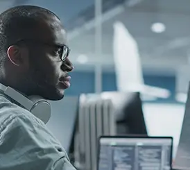 A man wearing glasses and headphones sits at a desk, looking at a computer screen with code displayed in an office setting.