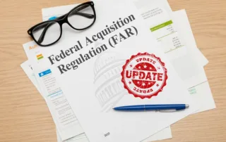 FAR-Update | Revolutionary Solutions A stack of papers labeled "Federal Acquisition Regulation (FAR)" with a red "UPDATE" stamp, a pair of glasses, and a blue pen on a wooden desk.